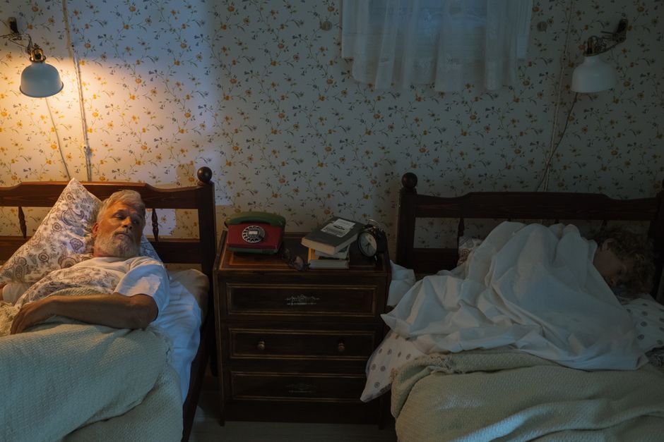 An elderly man and woman sleeping in a cozy bedroom at night, under warm light.